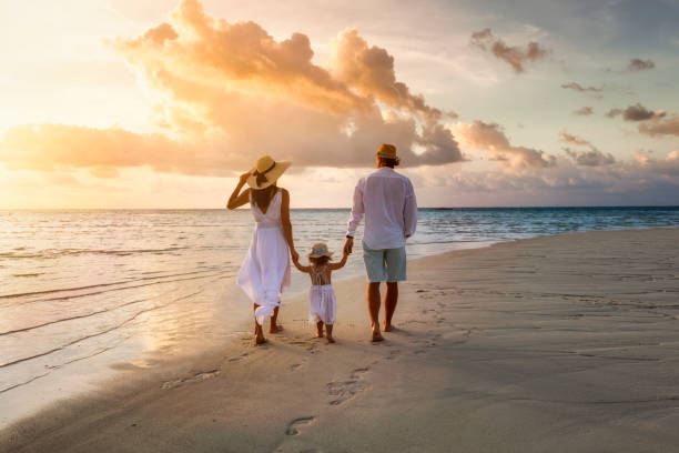 A elegant family in white summer clothing walks hand in hand down a tropical paradise beach during sunset tme and enjoys their vacation time