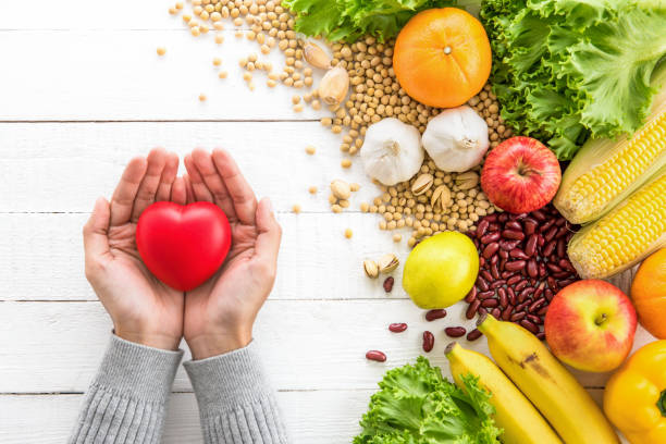 Woman hands holding red heart shape ball with various kinds of colorful healthy medicinal fruits, vegetables and nuts aside on white wood table